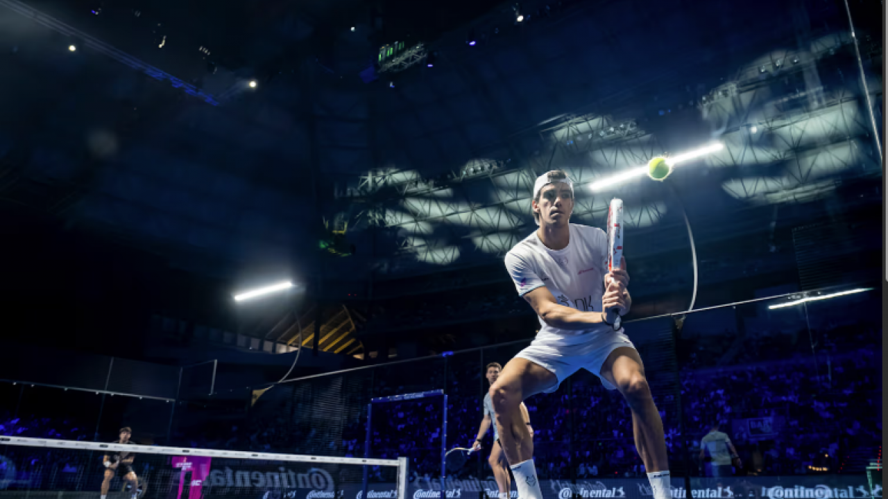 A professional padel player prepares for a fast volley during a high-intensity match inside a large indoor arena with dramatic lighting and a full crowd.
